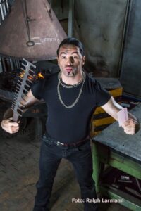 Bülent mit Kamm in der einen und Sägeblatt in der anderen Hand in einer Schmiede